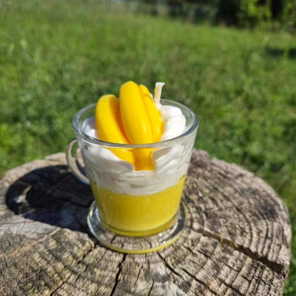 Bougie parfumée artisanale à la Banane dans une tasse en verre avec chantilly décorative blanche et petites bananes jaunes, posée en extérieur sur une souche en bois.