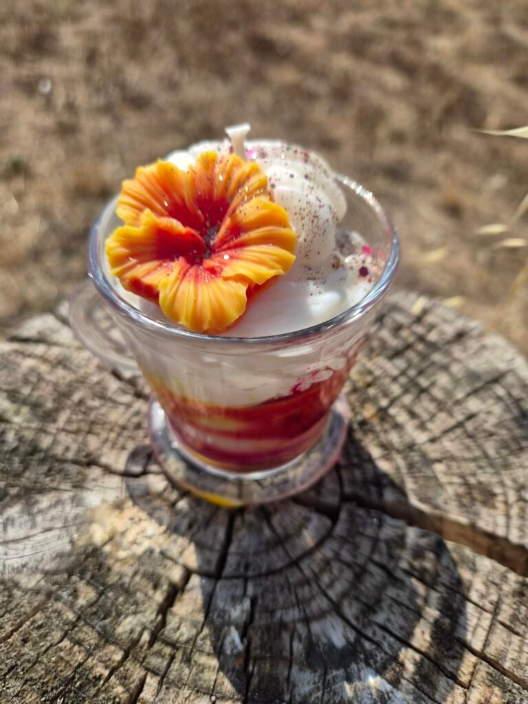 Bougie artisanale en cire de soja coulée dans une tasse en verre, avec des couches de cire rouge et blanche et une fleur orange décorative sur le dessus